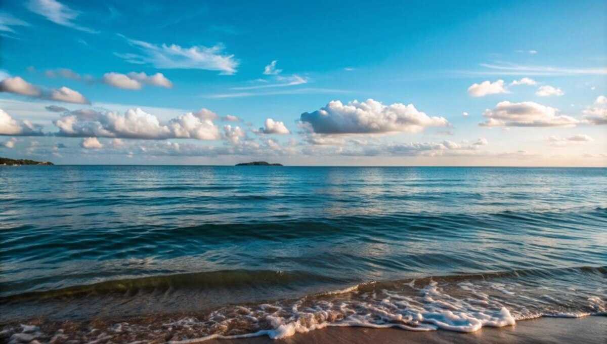 Calm blue ocean with gentle waves washing onto a sandy shore beneath a bright sky with white clouds